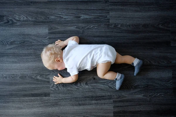 Upset baby in white bodysuit on the floor at home
