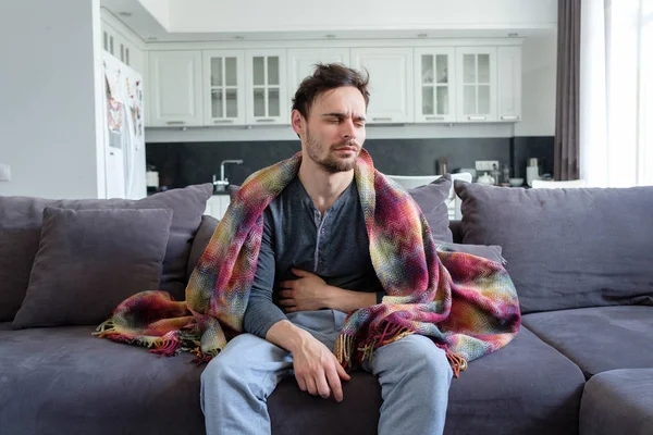 A sick man sits on a sofa in blankets and holds his stomach