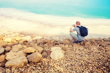 Dead sea alarak fotoğrafları cep telefonu ile genç adam. Kamera sahile geri üzerinde oturan adam
