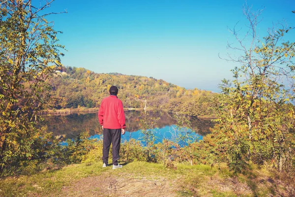A man stands on a hill above a beautiful forest lake on a sunny autumn day