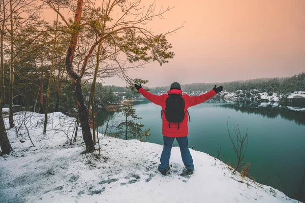 A man with hands in the air standing on the rocky lakeshore in winter. The man looking at the beautiful lake