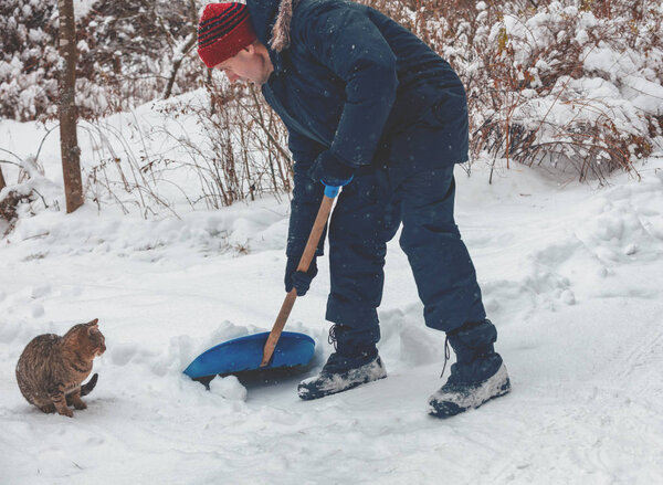 A man with shovel cleans the road from snow in snowfall. Cat sits nearby