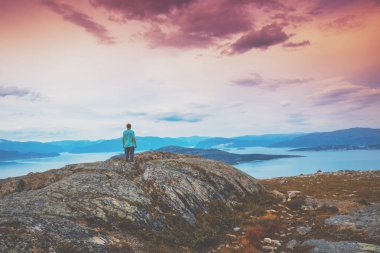 Fiyortun panoramik görüntüsü. Pembe gökyüzü ile alacakaranlık zamanı. Genç adam kayanın uçurumunda duruyor ve denize bakıyor. Gün batımında güzel bir dağ manzarası. İskandinav doğası, Norveç