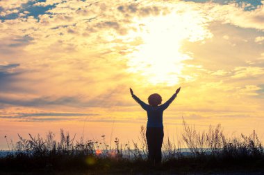 Happy young woman with hands in the air in a field during sunset. Silhouette of a happy young woman against the sunset sky. Girl looking at the sunset