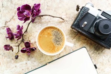 Orkideli bir fincan kahve, eski model analog kamera, mermer masanın üzerinde düz not defteri.