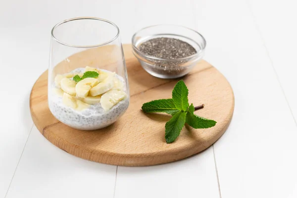Chia pudding with banana in a glass cup with a sprig of mint and grains in a bowl on a wooden cutting board on a white background. Copy space. A delicious snack consists of yogurt and grains. Horizontal orientation.