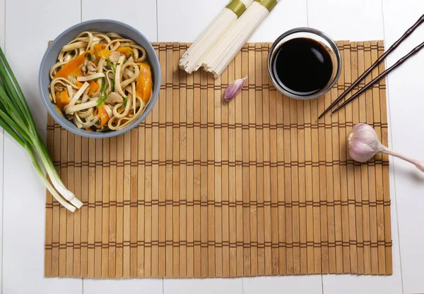 Udon noodles with chicken, soy sauce, carrots and fresh ingredients on a bamboo mat on a white background. Top view. Japanese kitchen. Copy space.