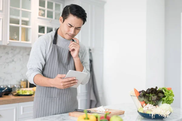 Asian confused man reading his tablet with a pensive thoughtful look while standing in his kitchen while cooking and preparing a meal from a variety of fresh vegetables on the counter in front of him