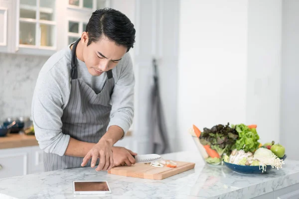 Asian confused man reading his tablet with a pensive thoughtful look while standing in his kitchen while cooking and preparing a meal from a variety of fresh vegetables on the counter in front of him