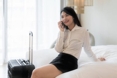 Güzel Asyalı genç gülümseyen işkadını kahve içme ve otel odasında kanepede otururken cep telefonuyla takım elbiseli. İş seyahati