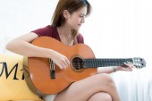 Mladá krásná Asijské žena sedící na gauči hraje na kytaru - girl hudba, baví precizní