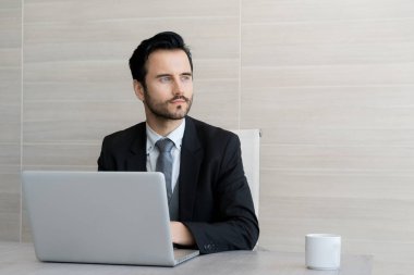 Yakışıklı işadamı bir dizüstü bilgisayar kullanarak, uzak bakıyor ve ofiste tabloda otururken gülümseyen.