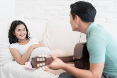 Güzel Asyalı hamile kadın ve Asya yakışıklı kocam gülümseyen ve evde yatakta akustik gitar çalmak.