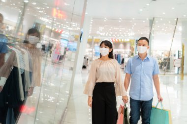 Yüzlerine maske takan Asyalı çift, alışveriş merkezinde alışveriş torbası takmış, Coronavirus 'u önleyen yeni bir elbise arıyor. Covid19 gribi.