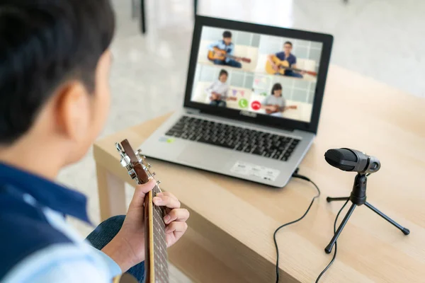 Asian boy playing acoustic guitar virtual happy hour meeting for play music online together with friend in video conference with laptop for a online meeting in video call for social distancing.
