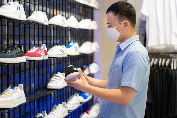 Asian man wearing mask over her face while choosing sport shoe for run at shopping mall for healthcare and prevention from coronavirus, Covid19 influenza in crowded place