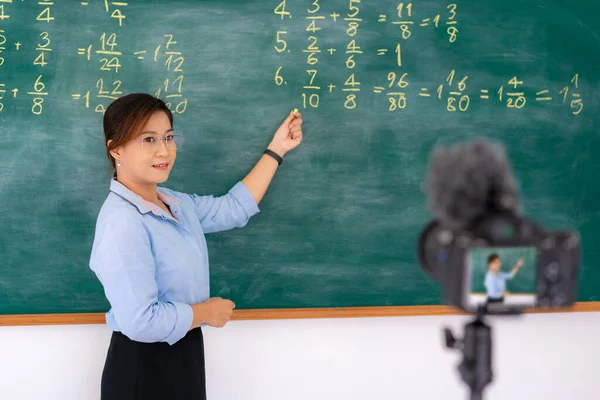 Asian primary teacher tutor explaining math in blackboard giving remote school class online lesson teaching looking at camera in classroom by e-learning livestream  video conference