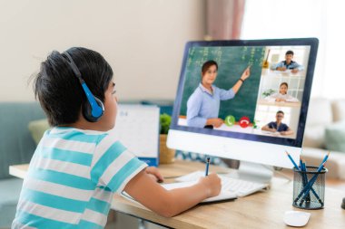 Asyalı çocuk öğrenci video konferansı öğretmenle ve sınıf arkadaşlarıyla evdeki oturma odasında bilgisayar başında. Evde eğitim ve uzak öğrenim, çevrimiçi, eğitim ve İnternet.