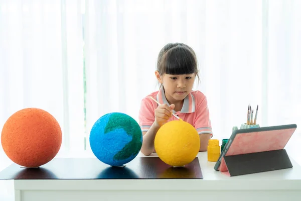 Asian elementary schoolgirl painting the moon in science class learning about the solar system via video conference with teacher and other classmates at home, Homeschooling and distance learning