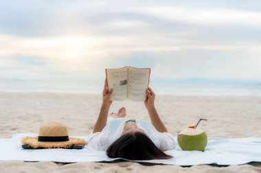  Genç Asyalı kadın, tropik bir yaz plaj denizinde uzanıp yanında hindistan cevizi suyuyla kitap okuyarak rahatlıyor. Yaz, tatil, tatil ve Tayland konseptinde mutlu insanlar..