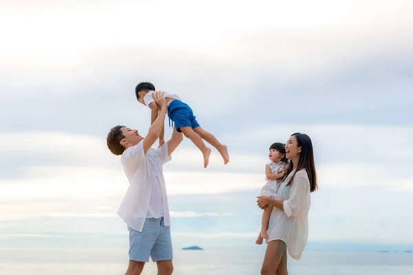 Happy Asian family holidays during joyful father, mother, son and daughter walking and raise son to sky along summer sunset sea. Happy family travel on beach in holiday, Summer and vacation.