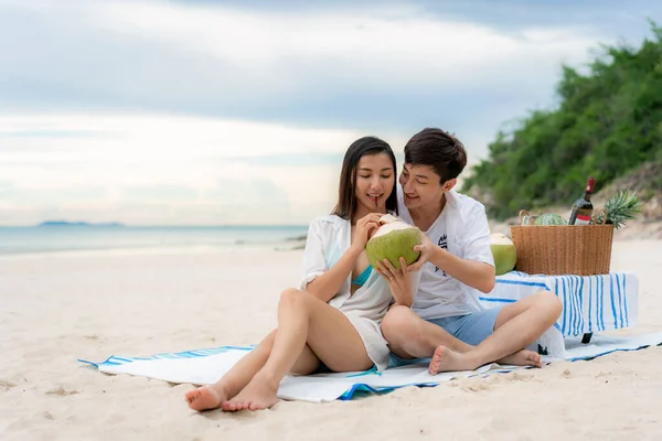 アジアの若いカップルがピクニック毛布の上に座って ビーチや海の近くでココナッツ水を飲みながら トロピカルフルーツを背景にしています タイの夏 幸せな人々のコンセプト — ストック写真