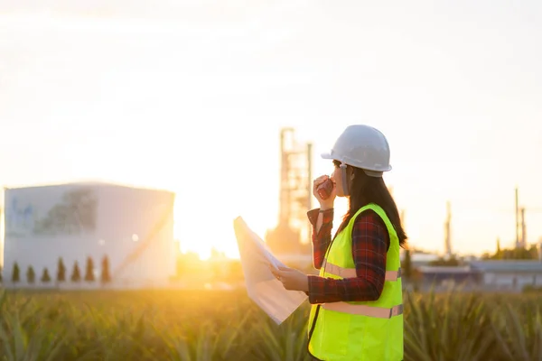 Asian woman technician Industrial engineer using walkie-talkie and holding bluprint working in oil refinery for building site survey in civil engineering project.