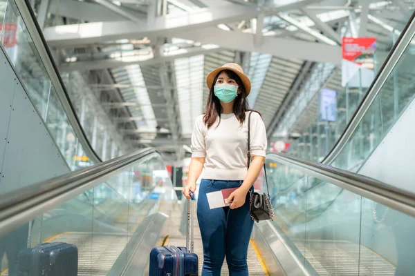エスカレーターの上に立つ空港のターミナルの外を見て顔マスクを着て荷物を持つアジアの旅行者の女性の旅のためのゲートに移動します 新常態旅行の概念 — ストック写真