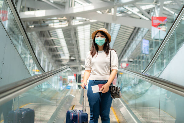 Asian traveler  woman with luggage wearing face mask looking outside terminal in airport standing on escalator go to gate for journey. New Normal travel concept.