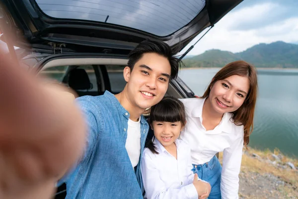 Portrait of Asian family sitting in car with father, mother and daughter selfie with lake and mountain view by smrtphone while vacation together in holiday. Happy family time.