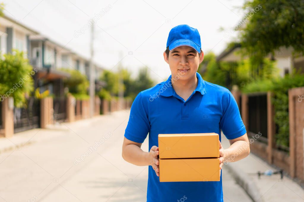 Asian delivery young man in blue uniform smile and holding pile of cardboard boxes in front house village with copy space. Advertising, Business, Transportation Concept