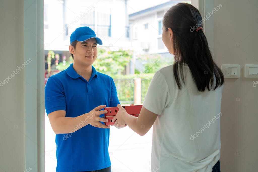 Asian delivery young man in blue uniform smile and holding pizza boxes in front house and Asian woman accepting a delivery of pizza boxes from deliveryman. Advertising, Business, Transportation.