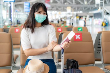 Maske takan Asyalı kadın turist, maskenin arkasında gülümserken mutlu görünüyor ve COVID-19 'un yeni normal seyahat konsepti korunurken havaalanı terminalinde sosyal uzaklık sandalyesinde oturuyor..