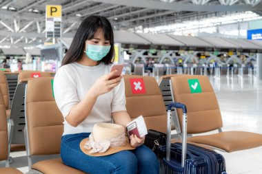 Havayolu uçuş statüsünü cep telefonuyla arayan maskeli bir Asyalı kadın turist ve COVID-19 'un yeni normal seyahat konsepti korunması sırasında havaalanı terminalinde sosyal mesafeli koltukta oturuyor..