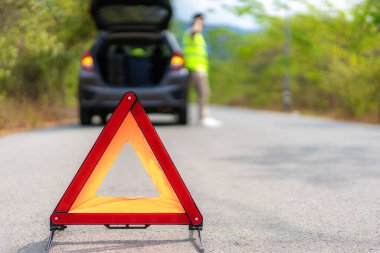 Breakdown triangle sign on road with worried Asian man talking by mobile phone with insurance or car service center company after car breakdown on country road in background clipart
