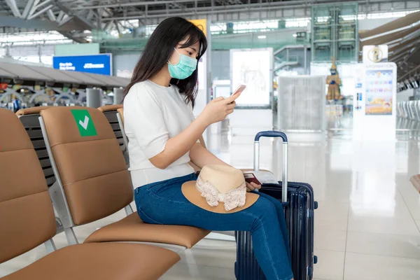 携帯電話の検索を使用してマスクを身に着けているアジアの女性観光客航空会社のフライトステータスと新しい通常の旅行コンセプトのためのCovid 19の保護中に空港ターミナルで社会的距離椅子に座って — ストック写真