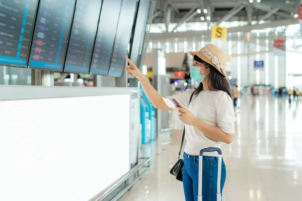 新しい通常の旅行のコンセプトのための空港ターミナルで到着出発ボードからのフライトをチェック顔マスクを身に着けているアジアの女性観光客 — ストック写真