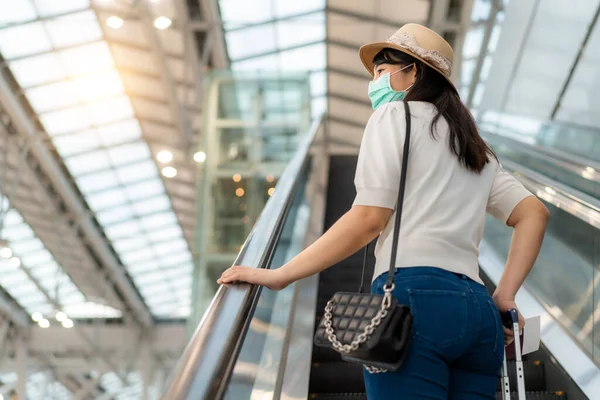 エスカレーターの上に立つ空港のターミナルの外を見て顔マスクを着て荷物を持つアジアの旅行者の女性の旅のためのゲートに移動します 新常態旅行の概念 — ストック写真