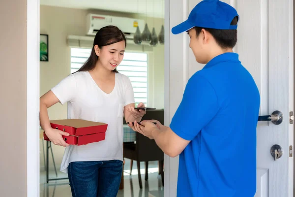 Asian delivery young man in blue uniform smile and holding food boxes in front house and Asian woman accepting a delivery of food boxes and payment QR code by smartphone from deliveryman.