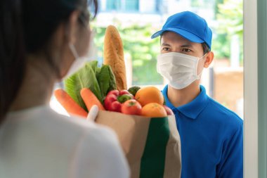 Asyalı teslimatçı, maske ve eldiven takıyor. Yiyecek, meyve ve sebzelerle dolu bir çanta. Ev izolasyonu sırasında kadın kostümcüyü evin önünde bekliyor..
