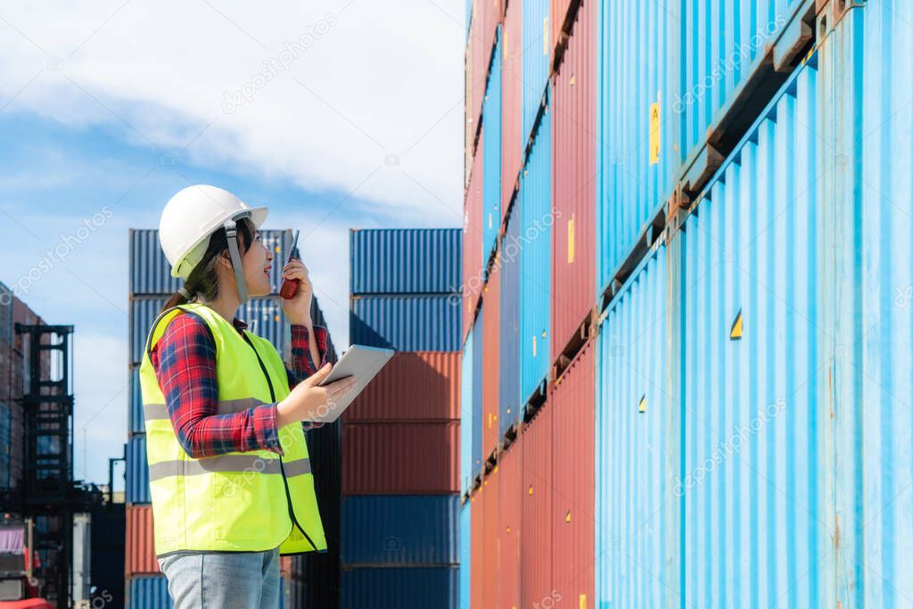 Engineer woman or Foreman manager in container depot working with digital tablet and walkie talkie for control and checking industrial container yard for Logistics oversea import export shipping business in port