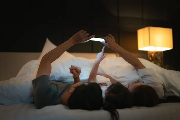 Asian happy family mother, father and little daughter watching movie or cartoon in smartphone together and blanket cover their head in bed at night at home