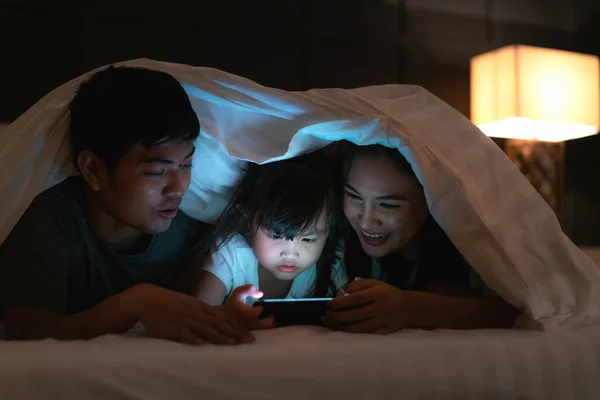 Asian happy family mother, father and little daughter watching movie or cartoon in smartphone together and blanket cover their head in bed at night at home
