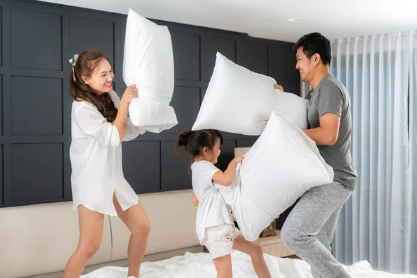 Asian happy family having funny pillow fight on white bed in bedroom for free time with father, mother and daughters feeling happy and family relationship in bedroom at home