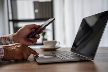 Konsept ile iletişime geçin: dizüstü bilgisayarın yanında akıllı telefon kullanan eller, modern çalışma alanında parmakla dokunmak; müşteri desteği ve soruşturma iletişimi, yardım masası servisi, cep telefonu mesajlaşması, iş bağlantısı.