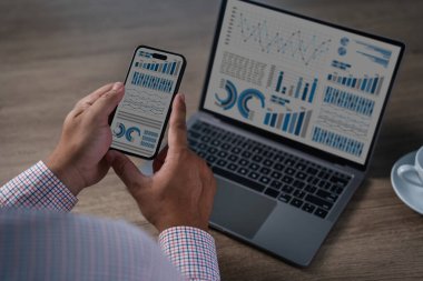 Modern bir çalışma alanında grafikleri ve grafikleri gösteren akıllı telefonu kontrol eden eller; iş performansı, satış, pazarlama, finans, gerçek zamanlı içgörü için veri, ölçüm ve raporlar.