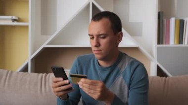 Kanepede oturan genç adam, kredi kartıyla online alışveriş yapmak için akıllı telefon kullanıyor. Kapalı.