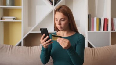 Kredi kartı ve akıllı telefon tutan genç bayan müşteri evdeki kanepede oturuyor. İnternetten alışveriş yapan mutlu bayan alışverişçi kolay taşınabilir ödemeler yapıyor. E-bankacılık uygulama servisi