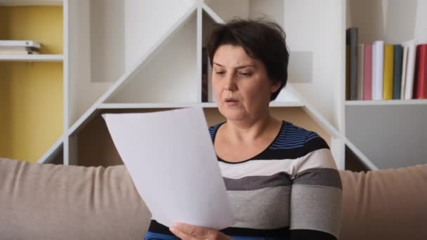 Traurige Frau, die zu Hause auf der Couch sitzt, liest empfangene schlechte Nachrichten, hält Dokumente auf Papier und fühlt sich über finanzielle Probleme verzweifelt. Ausweisungskonzept — Stockvideo