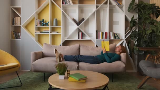 Fiatal nő fekszik a kanapén otthon. elgondolkozva és álmodozva olvasni egy könyvet. nyugodt és kényelmes — Stock videók